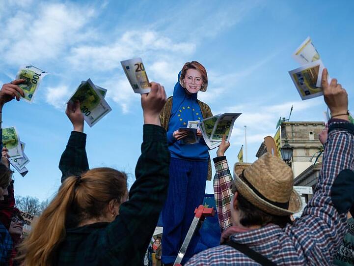 Bei einer Aktion am Rande der Demonstration verteilt eine Aktivistin mit einer Von-der-Leyen-Maske symbolisch nachgemachte Geldscheine.