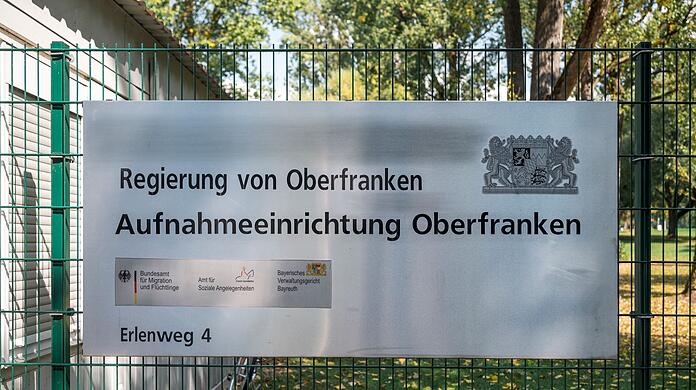 Ein Hinweisschild der Aufnahmeeinrichtung Oberfranken hängt am Zaun der Flüchtlingsunterkunft.