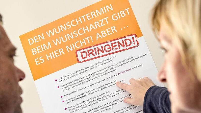 Ein Mann und eine Frau betrachten ein Plakat mit Hinweisen zu der Einführung von "Terminservicestellen".