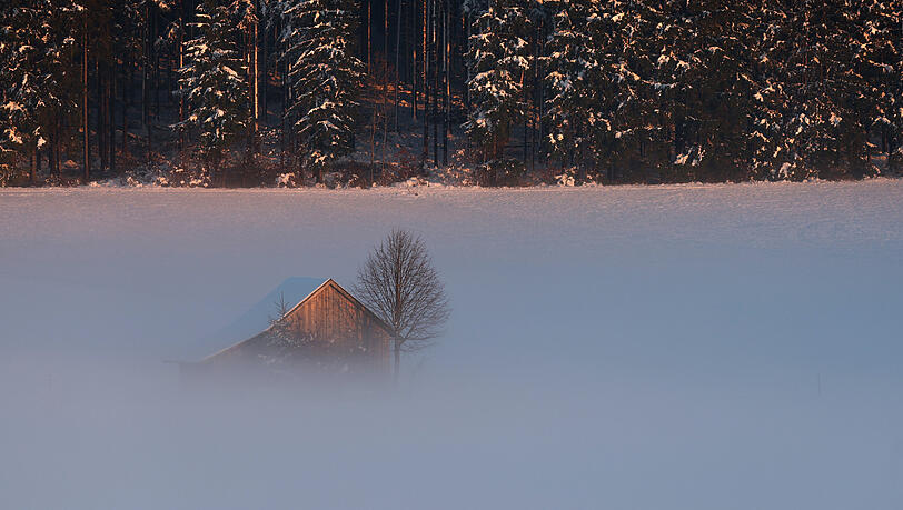 Eine Hütte in der im Abendnebel liegenden Schneelandschaft wird von der untergehenden Sonne beschienen. Eine Hütte in der im Abendnebel liegenden Schneelandschaft wird von der untergehenden Sonne beschienen.