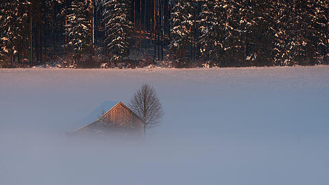 Eine Hütte in der im Abendnebel liegenden Schneelandschaft wird von der untergehenden Sonne beschienen.