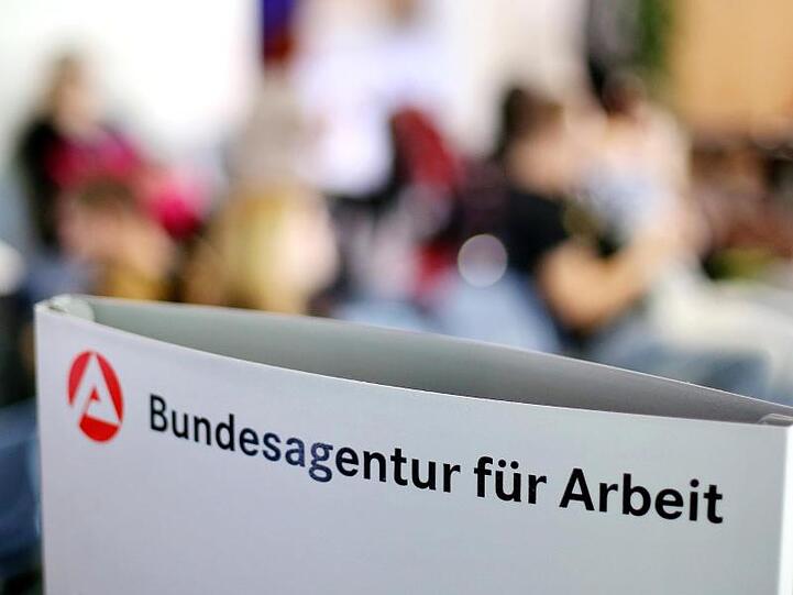 Ein Schild mit der Aufschrift "Bundesagentur für Arbeit" steht einem vollen Wartezimmer im Jobcenter.