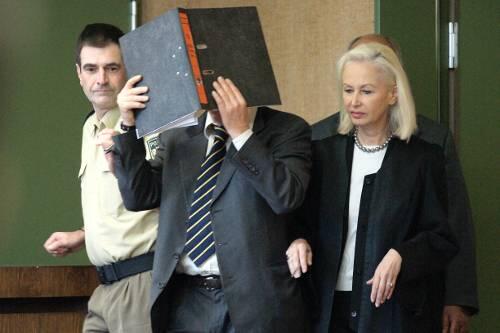 Der Angeklagte betritt den Gerichtssaal mit einem Aktenordner vor dem Gesicht,