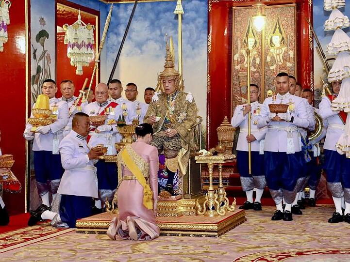 Thailands König Maha Vajiralongkorn (M) auf dem Thron, vor ihm kniet seine Frau, Königin Suthida.