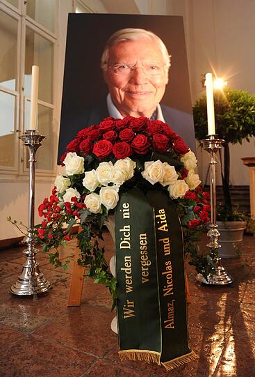 Ein Gesteck mit roten und weißen Rosen und Schleifen mit dem Text "Wir werden Dich nie vergessen - Almaz, Nicolas und Aida" steht vor einem Porträt von Karlheinz Böhm.