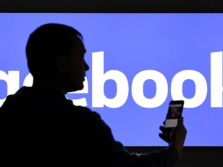 Ein Mann mit Smartphone steht vor einem Monitor mit dem Logo von Facebook.