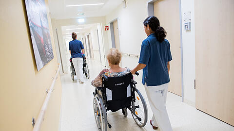 Zwei Pflegerinnen schieben in einem Klinikum Patienten mit Rollstühlen durch einen Flur.