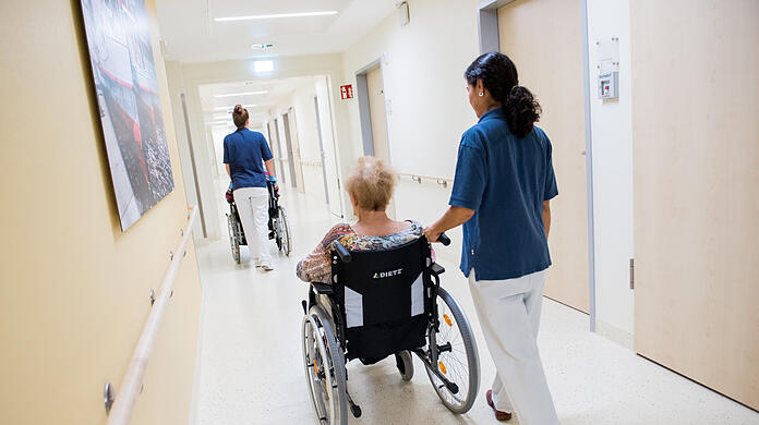Zwei Pflegerinnen schieben in einem Klinikum Patienten mit Rollstühlen durch einen Flur.