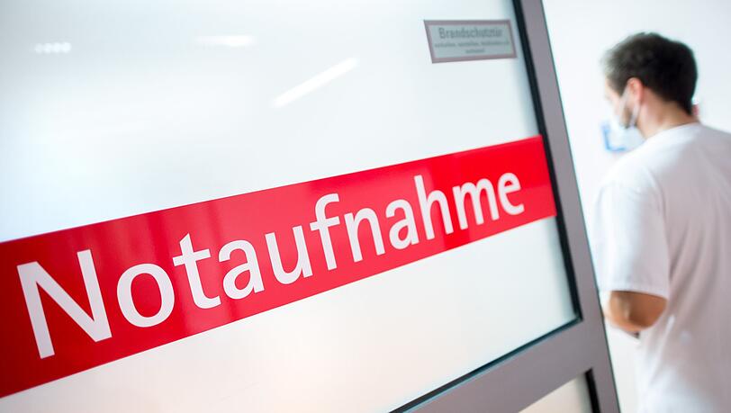Ein Krankenpfleger steht in einem Krankenhaus neben einer Glastür mit der Aufschrift "Notaufnahme".