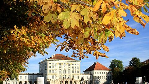 Passanten und Spaziergänger geniessen die warmen Sonnenstrahlen und das bunte Herbstlaub bei einem Spaziergang oder sitzen auf einer Parkbank in der Sonne im Schlosspark Nymphenburg in  München