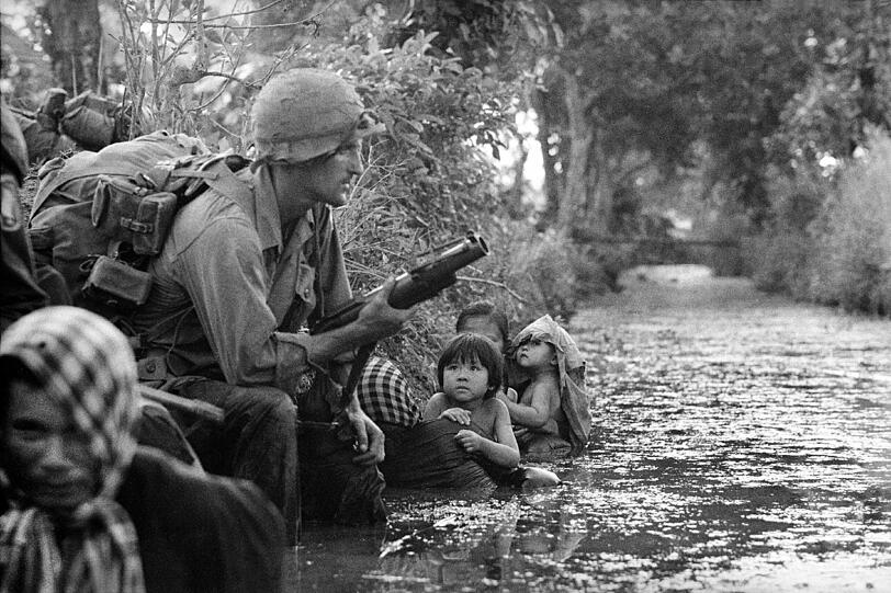 Zwei südvietnamesische Kinder starren einen amerikanischen Fallschirmjäger an, der einen M79 Granatenwerfer hält. Sie halten sich an ihren Müttern fest, die sich an einem Flussufer festklammern, um sich vor Scharfschützen der Vietcong in Sicherheit zu bringen. Bao Trai, 20 Meilen westlich von Saigon, 1966.
