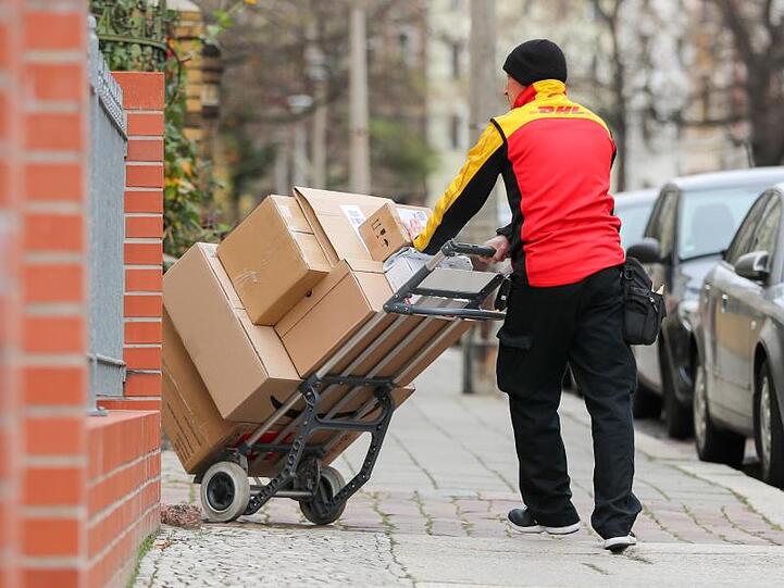 Ein DHL-Paketzusteller geht mit einer Sackkarre voller Pakete zu einem Haus.