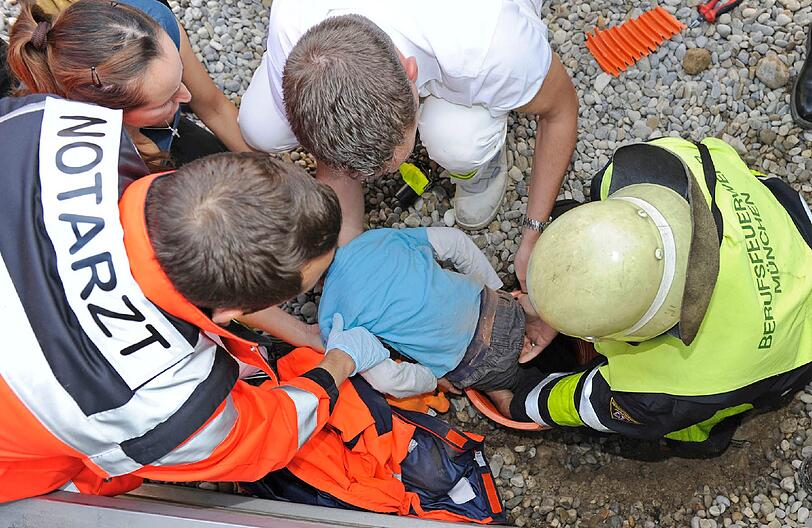 Mit vereinten Kräften wird das Kind gerettet: Ein Feuerwehrmann schaufelt das Rohr frei, einer hält eine Zange zum Aufschneiden in der Hand, wieder andere beruhigen den Kleinen. Auch die Mutter spricht beruhigend auf ihren Sohn ein.
