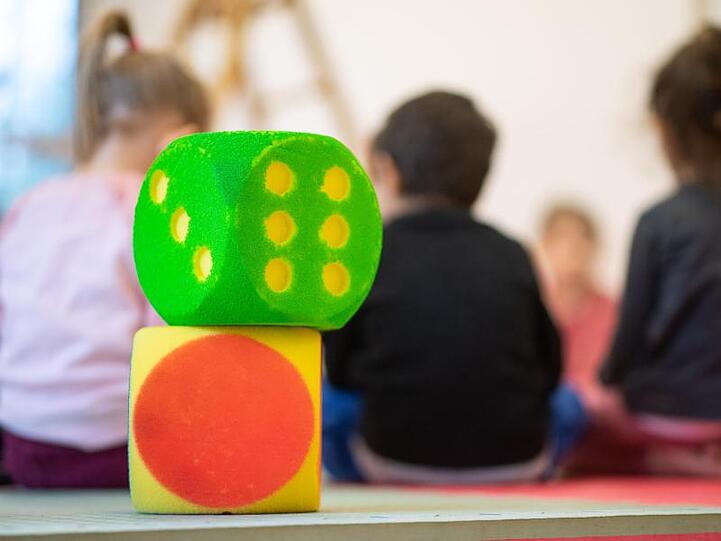 Zwei Würfel stehen in einer Kindertagesstätte vor Kindern, die in einem Kreis sitzen. Foto: Sebastian Gollnow/Archivbild