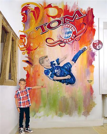 Der junge Tom Bischof unter dem Wandgemälde, das ihn und das Logo des FC Bayern zeigt.