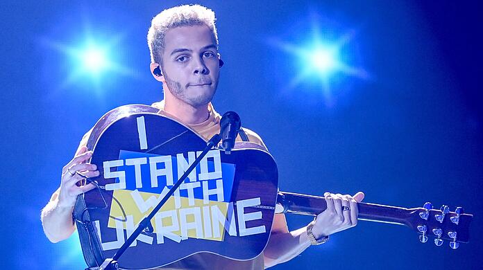 Malik Harris steht bei der Show "Germany 12 Points - der deutsche ESC-Vorentscheid" auf der Bühne und zeigt seine Gitarre, auf der "I stand with Ukraine" steht.