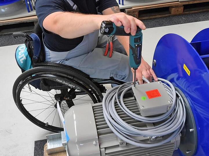 An einem Arbeitsplatz für Motorklemmen ist ein Mitarbeiter im Rollstuhl mit Montagearbeiten beschäftigt.