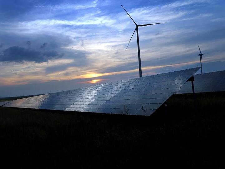 Der mit Wolken überzogene Abendhimmel spiegelt sich auf den Solarzellen einer vor Windrädern stehenden Solarkraftanlage in Kitzingen. Foto: Karl-Josef Hildenbrand/dpa/Archiv