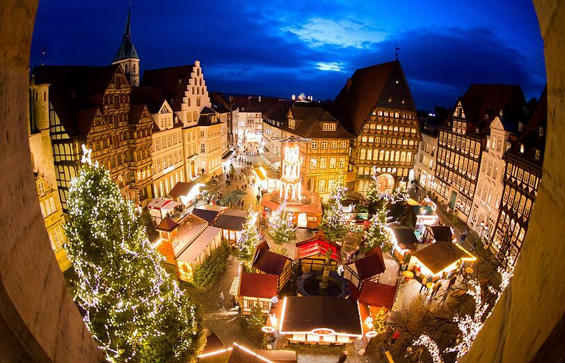 Festlich geschmückte Stände, Weihnachtsbäume und Lichter illuminieren zur "blauen Stunde" den Weihnachtsmarkt auf dem historischen Marktplatz in Hildesheim. Auf dem Marktplatz ist mittig mit Spitzgiebel das Knochenhaueramtshaus zu sehen.