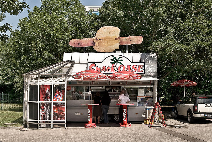 Das ist mal ein subtiler Hungermacher: Wer nach dem Rewe-Einkauf in Ismaning noch nicht ahnt, dass sein Magen eine Bratwurst braucht, entdeckt es spätestens am Parkplatz - beim Blick auf die Riesenwurscht auf dem Dach der "Snack Oase". "Den Imbiss hat es schon gegeben, als ich in den 90er-Jahren zur Schule gegangen bin", erinnert sich Fotograf Juri Gottschall. 2008 steht der rollende Imbiss unverändert da.