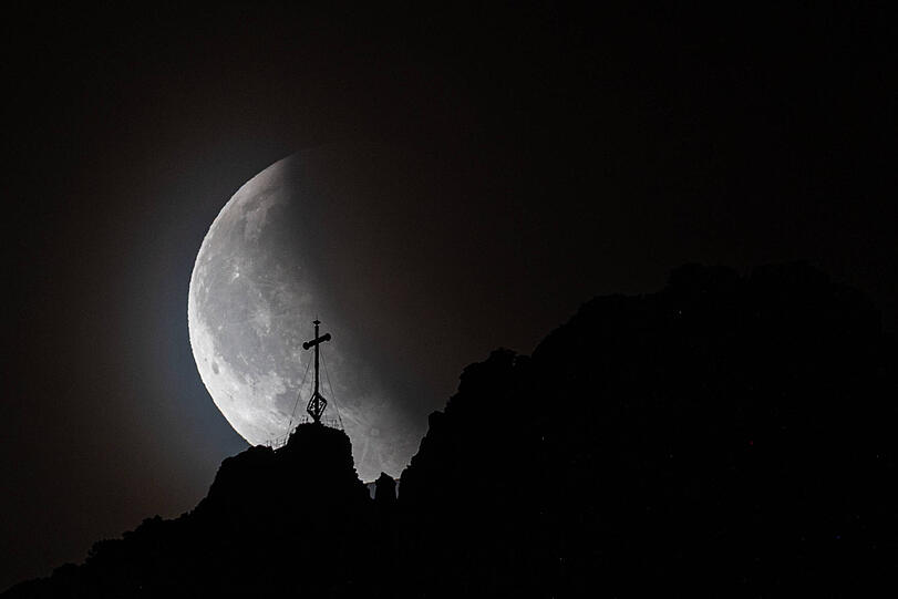 Mondfinsternis vor der Kampenwand. Der Vollmond ist während einer totalen Mondfinsternis vor dem Gipfelkreuz der Kampenwand in den bayerischen Alpen in Aschau zu sehen.