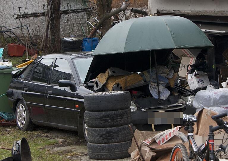 Auf dem Hof steht ein kaputtes Auto - mit Regenschirm über der Motorhaube.