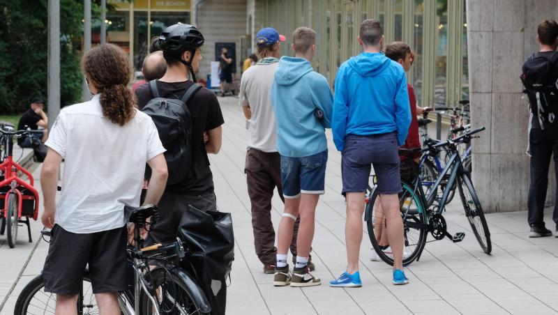 Kunden einer Fahrradwerkstatt stehen vor der Tür des Geschäftes in einer Schlange an.