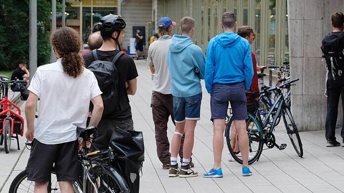 Kunden einer Fahrradwerkstatt stehen vor der Tür des Geschäftes in einer Schlange an.