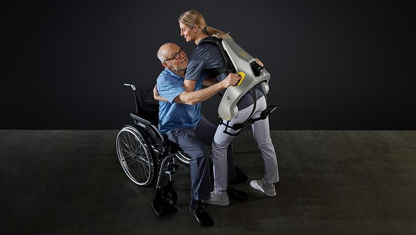 Eine Frau hebt mit Hilfe eines intelligenten Kraftanzugs der Firma German Bionic einen Mann aus einem Rollstuhl.
