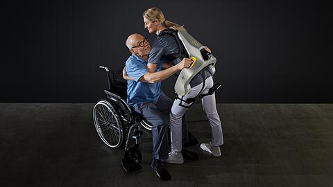 Eine Frau hebt mit Hilfe eines intelligenten Kraftanzugs der Firma German Bionic einen Mann aus einem Rollstuhl.