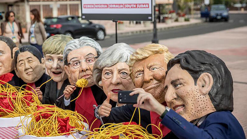 Aktivisten von Oxfam sitzen mit Masken der G7 Regierungschefs in Taormina an einem symbolisch gedeckten Tisch.
