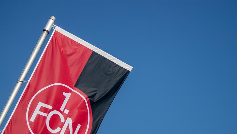 Eine Vereinsfahne mit dem Logo des 1. FC Nürnberg weht vor blauem Himmel.