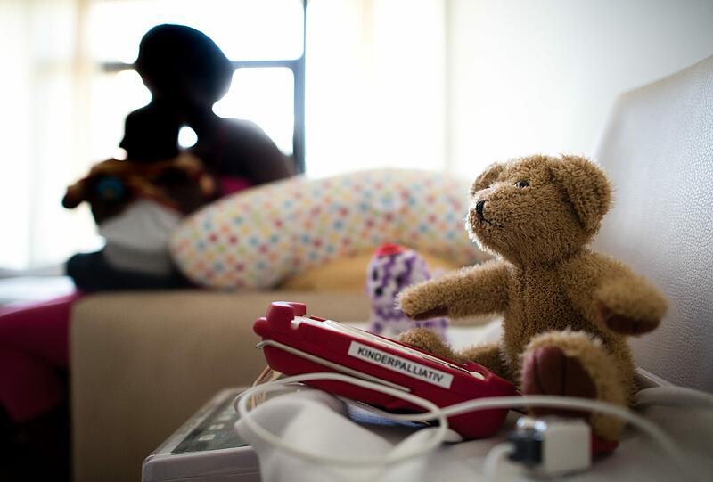 Eine Teddybär und ein Pulsoximeter in einem Patientenzimmer: Im Vordergrund eine Mutter, die ihren erkrankten Säugling im Arm hält.