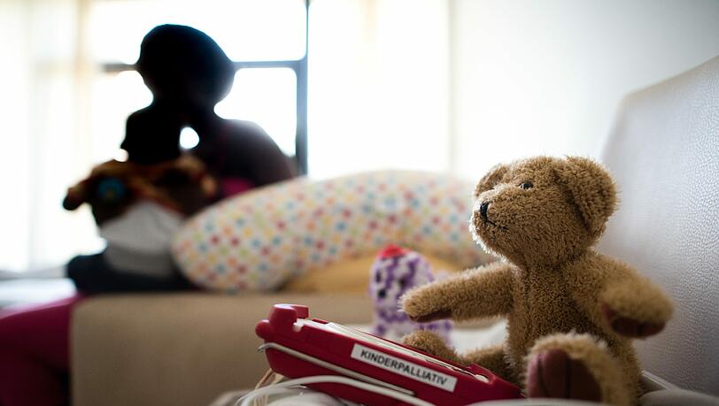 Eine Teddybär und ein Pulsoximeter in einem Patientenzimmer: Im Vordergrund eine Mutter, die ihren erkrankten Säugling im Arm hält. Eine Teddybär und ein Pulsoximeter in einem Patientenzimmer: Im Vordergrund eine Mutter, die ihren erkrankten Säugling im Arm hält.