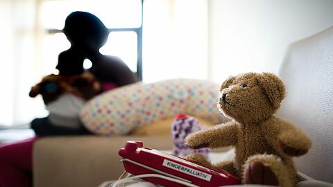Eine Teddybär und ein Pulsoximeter in einem Patientenzimmer: Im Vordergrund eine Mutter, die ihren erkrankten Säugling im Arm hält.
