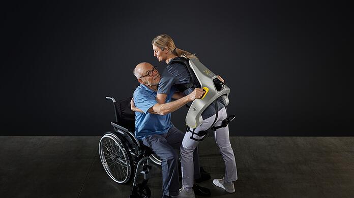 Eine Frau hebt mit Hilfe eines intelligenten Kraftanzugs der Firma German Bionic einen Mann aus einem Rollstuhl.