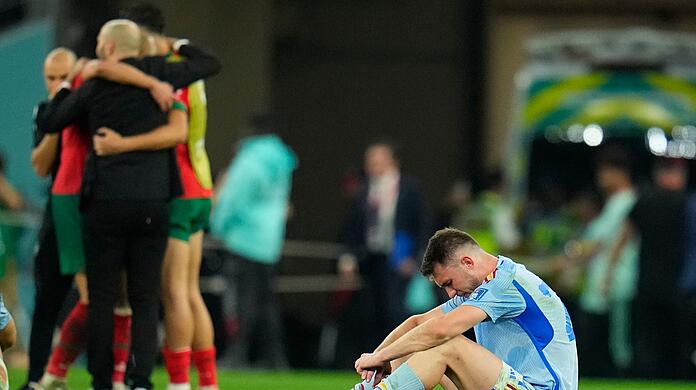 Spaniens Aymeric Laporte sitzt nach der Niederlage auf dem Spielfeld, während Marokkos Spieler im Hintergrund über ihren Sieg im Elfmeterschiessen jubeln.