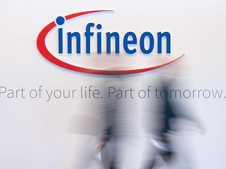 Menschen gehen vor einer Wand vorbei, die das Infineon-Logo zeigt. Foto: Lino Mirgeler/Archiv