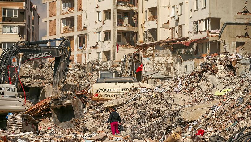 Ein Mann steht in Samandag in der Türkei zwischen Trümmern, während Bagger die Überreste eines zerstörten Gebäudes abtransportieren. Nach dem katastrophalen Beben am 6.2. kommt es in der Region immer wieder zu Nachbeben.