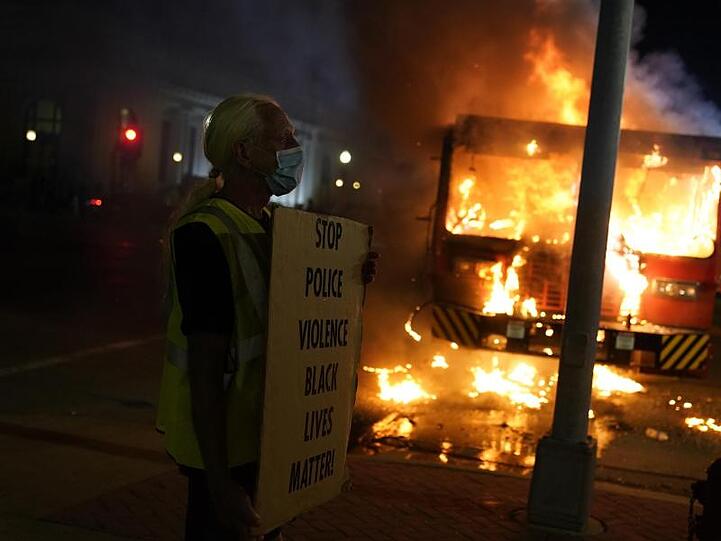 Ein Demonstrant steht mit einem Transparentvor einem brennenden Müllwagen.