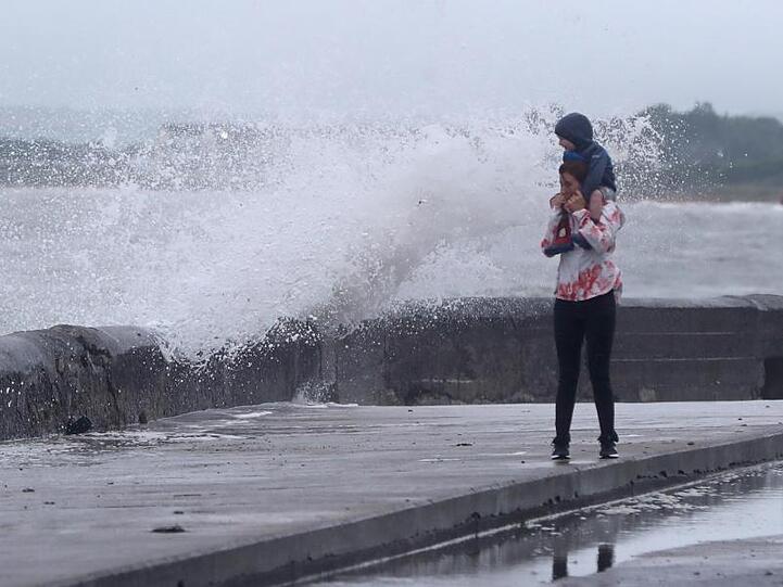 Eine Welle tritt über das Ufer während eine Frau mit einem Kind auf den Schultern an der Strandpromenade geht. Sturmtief "Ellen" trifft auf die Küste Irlands.