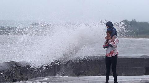 Eine Welle tritt über das Ufer während eine Frau mit einem Kind auf den Schultern an der Strandpromenade geht. Sturmtief "Ellen" trifft auf die Küste Irlands.