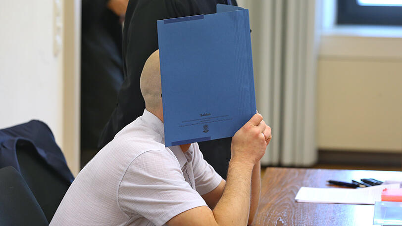 Der Angeklagte sitzt in einem Gerichtssaal im Landgericht auf der Anklagebank und verdeckt dabei mit einem Aktendeckel sein Gesicht.