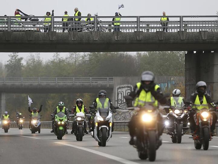 Demonstranten stehen auf einer Brücke, während Motorradfahrer mit gelben Westen auf der Straße unterwegs sind, um gegen die Spritpreise zu protestieren.