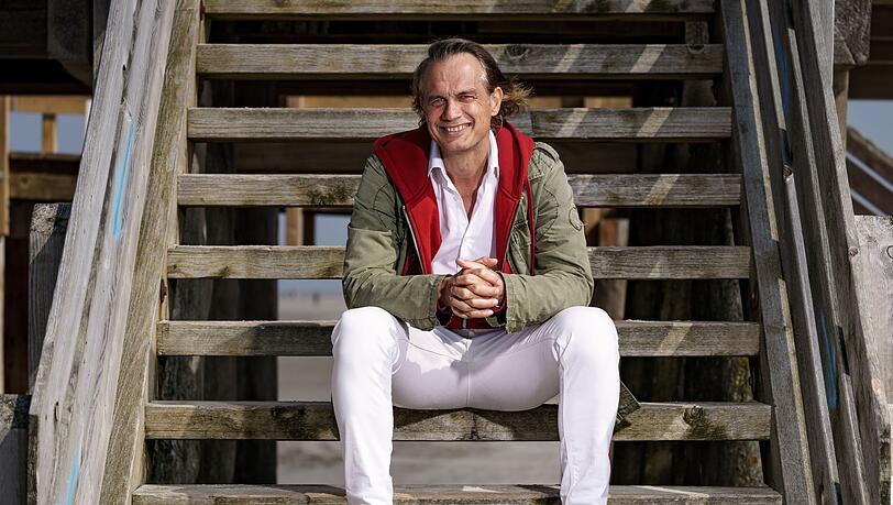 Schauspieler Ralf Bauer sitzt am Strand auf der Treppe eines der traditionellen Pfahlbauten.