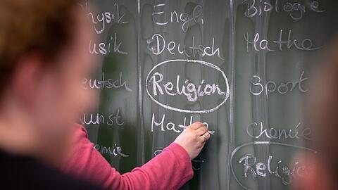 Eine Lehrerin schreibt mit Kreide einen Stundenplan mit Religionsunterricht an eine Tafel.