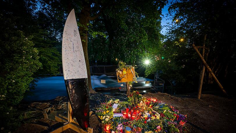 Blumen, Kerzen und Briefe liegen in den fr&uuml;hen Morgenstunden vor einem Zaun am Ufer der Eisbachwelle im Englischen Garten. Links ist ein Surfboard aufgebaut, auf dem Nachrichten und Unterschriften hinterlassen wurden. Eine Surferin, die im April auf der Eisbachwelle im Englischen Garten st&uuml;rzte, war im Krankenhaus verstorben.