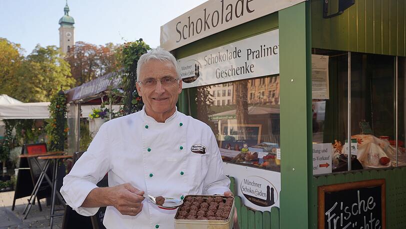 Konditormeister Josef Obermeier vor seinem Standl am Viktualienmarkt. Er hält seine Jubiläums-Pralinen zum 10-Jährigen hoch. Die sind bei der Kundschaft ein Renner.