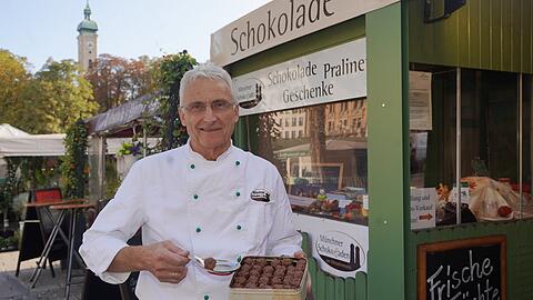 Konditormeister Josef Obermeier vor seinem Standl am Viktualienmarkt. Er hält seine Jubiläums-Pralinen zum 10-Jährigen hoch. Die sind bei der Kundschaft ein Renner.