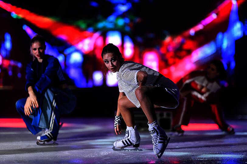 Drei Eiskunstläuferinnen zeigen vor einer bunten Lichtkulisse eine dynamische Performance